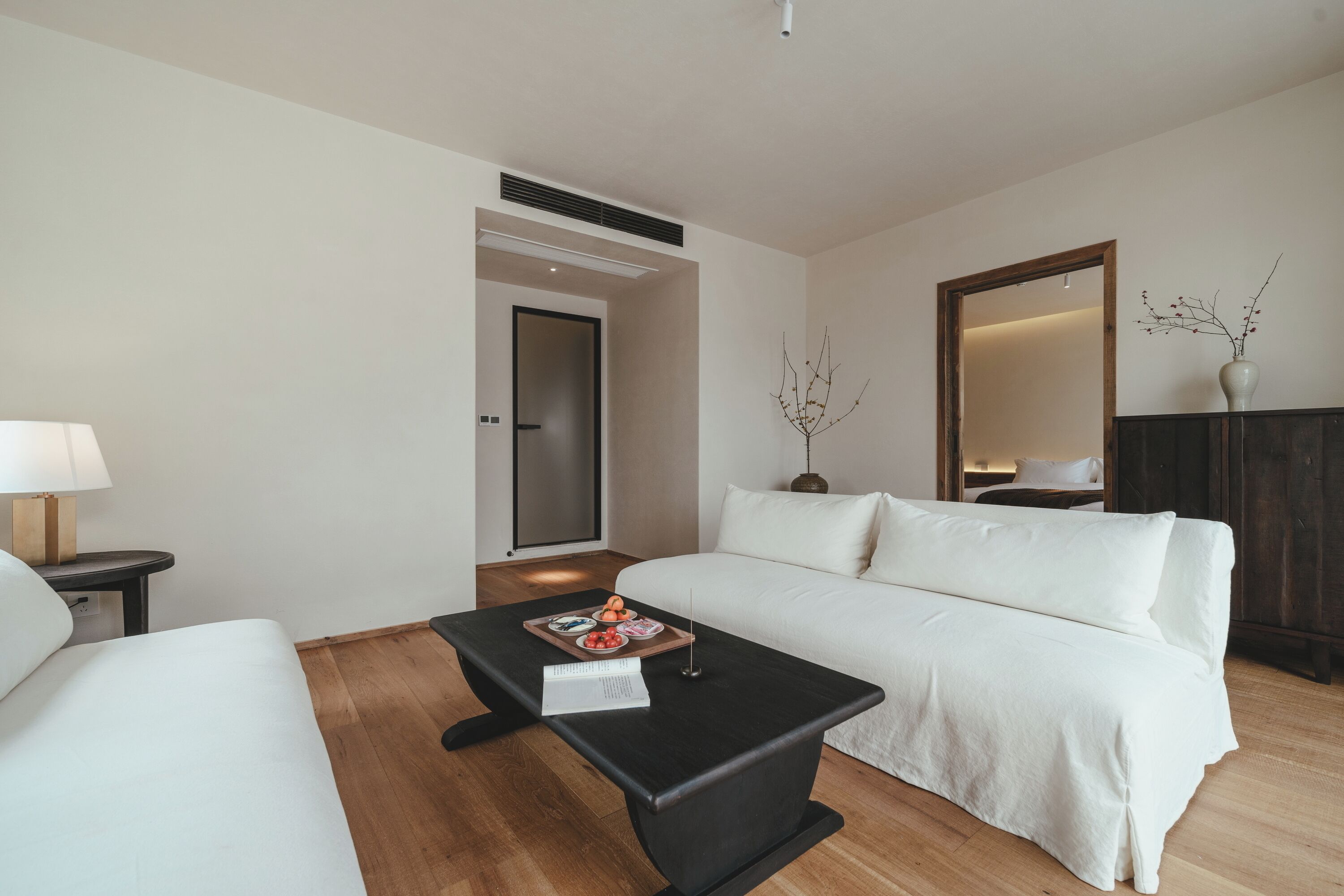 Una habitación de hotel moderna con un sofá blanco, una mesa de centro negra con una bandeja de aperitivos, una lámpara y un espejo grande.