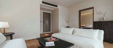 Una habitación de hotel moderna con un sofá blanco, una mesa de centro negra con una bandeja de aperitivos, una lámpara y un espejo grande.