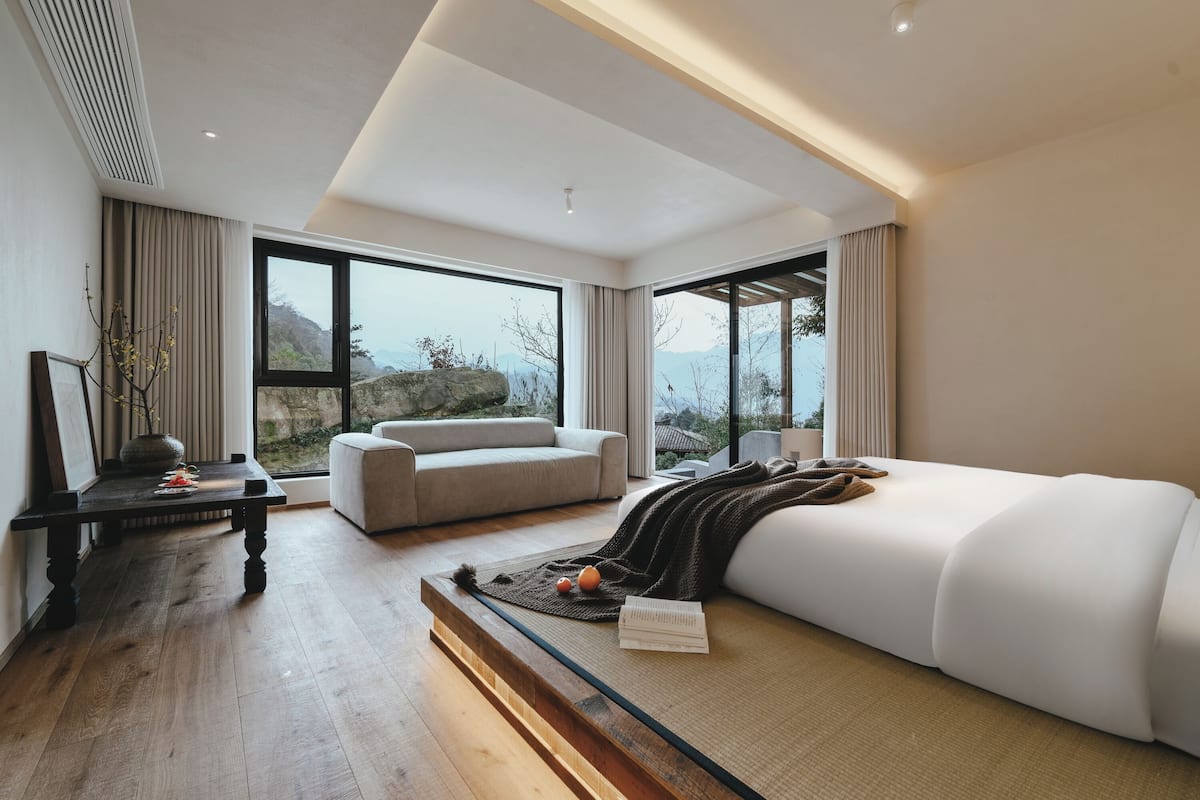 A modern hotel room with a large bed, a sofa, and a dining table. The room has large windows offering a view of the mountains.