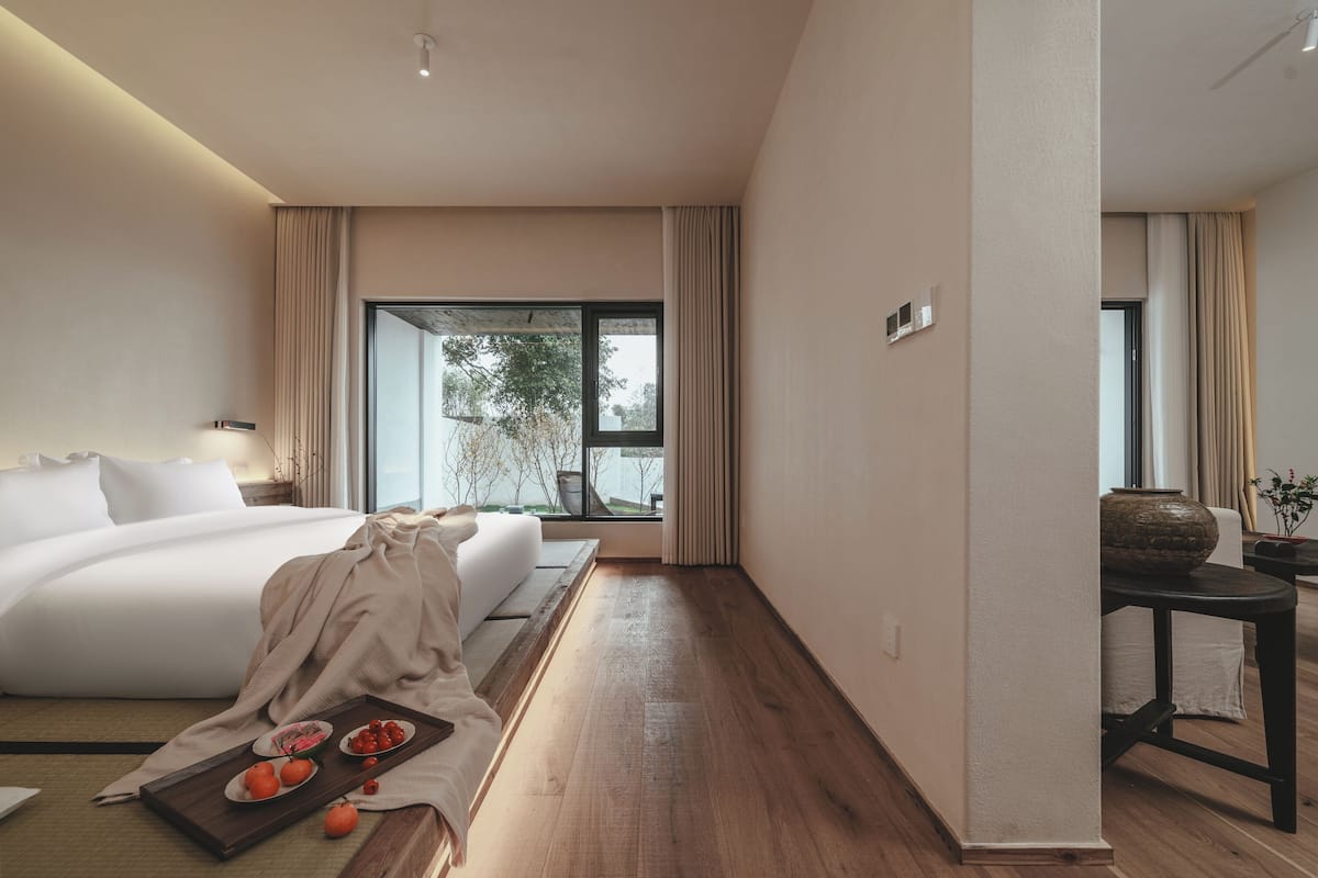 A modern hotel room with a large bed, a wooden floor, a dining table with a tray of fruit, and a view of trees through the window.