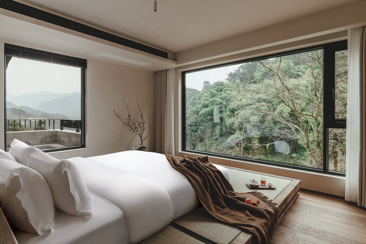 A bedroom with a large window, a bed, a bedside table with a cup and saucer, and a view of a forested area.