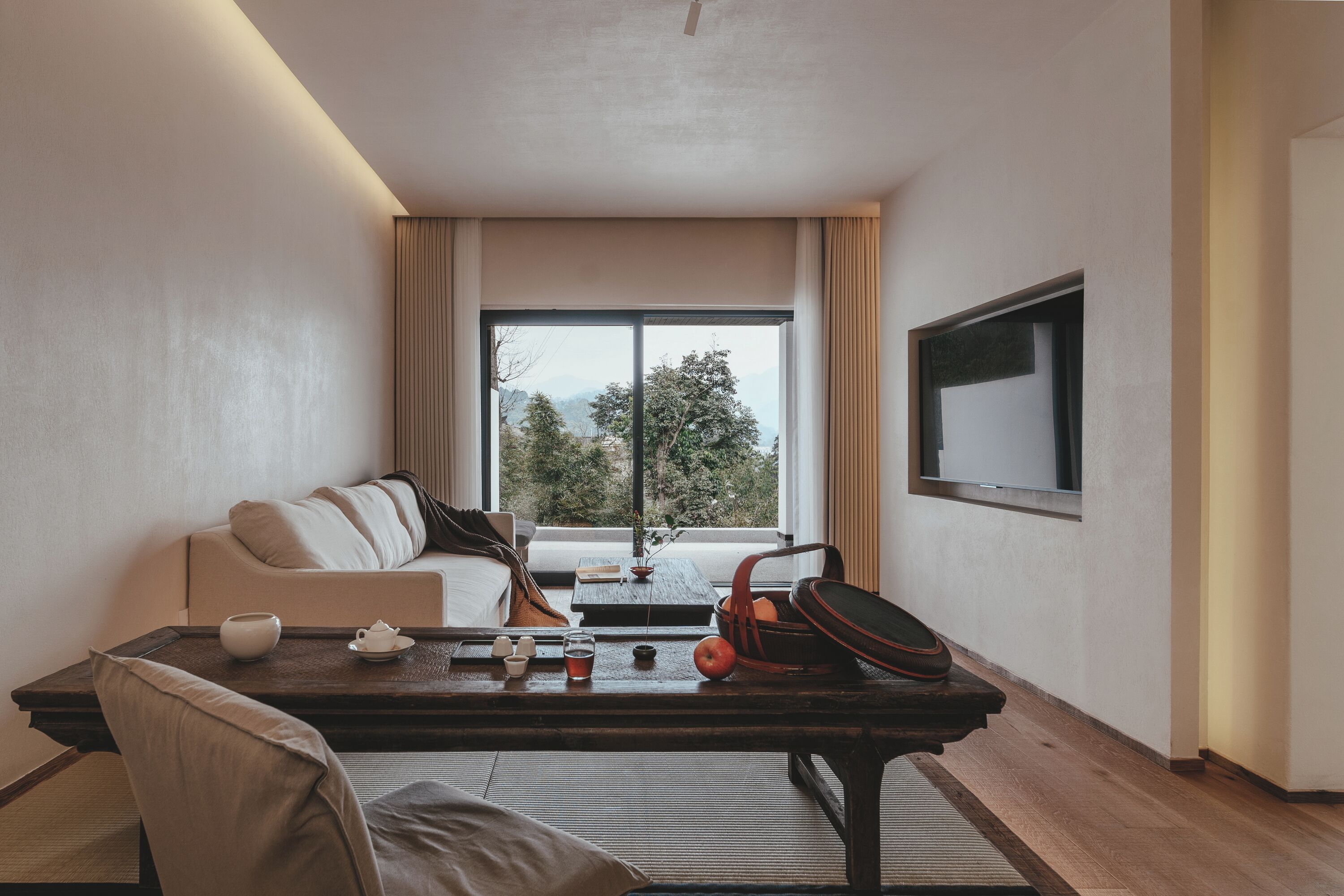 Una habitación de hotel moderna con un sofá, una mesa de madera con tetera, tazas y un vaso de agua, un televisor en la pared y un ventanal con vistas a árboles y montañas.