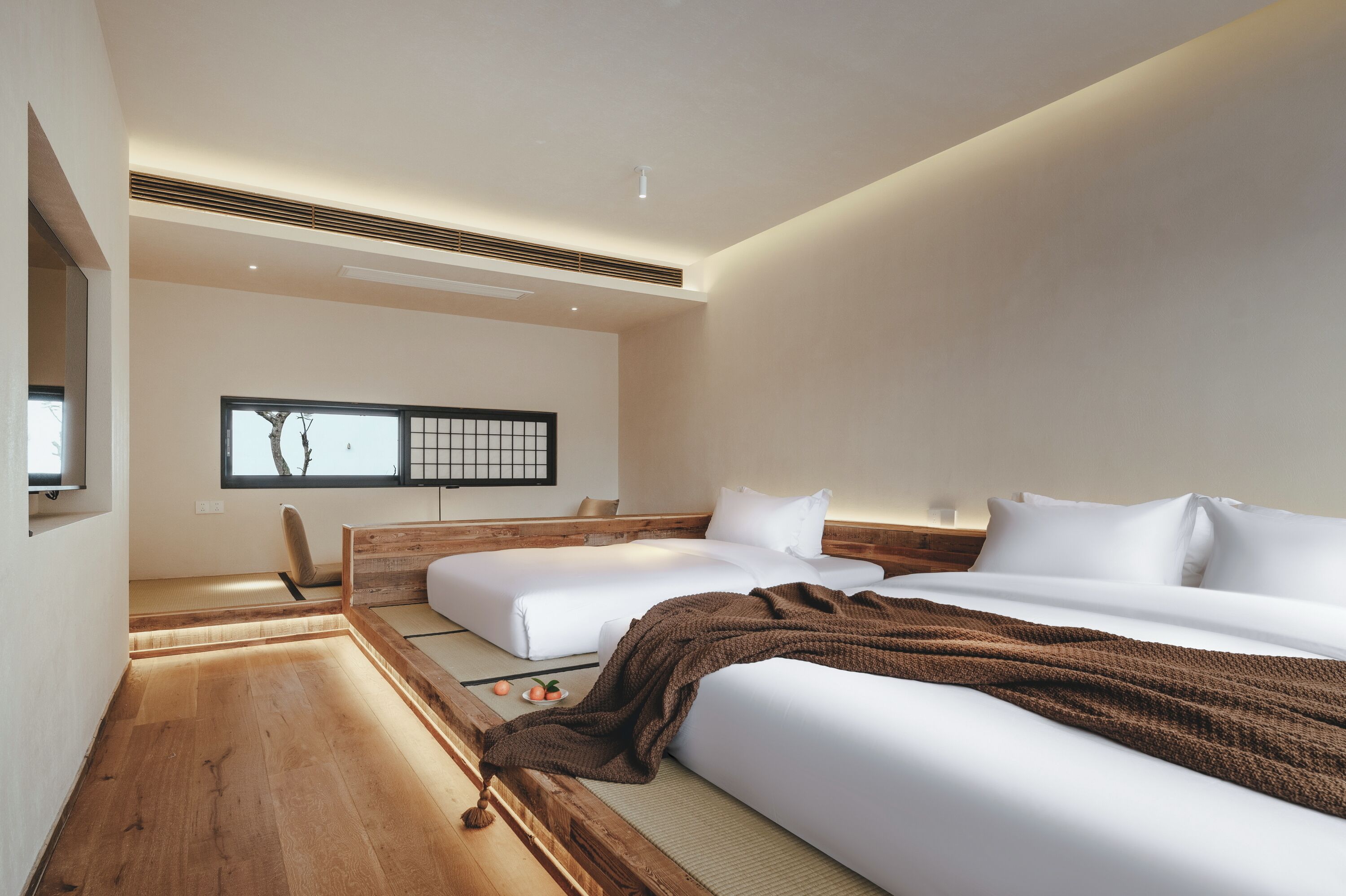 Quarto de hotel moderno com duas camas, piso de madeira, esteira de tatame, janela ampla com vista e televisão fixada na parede.