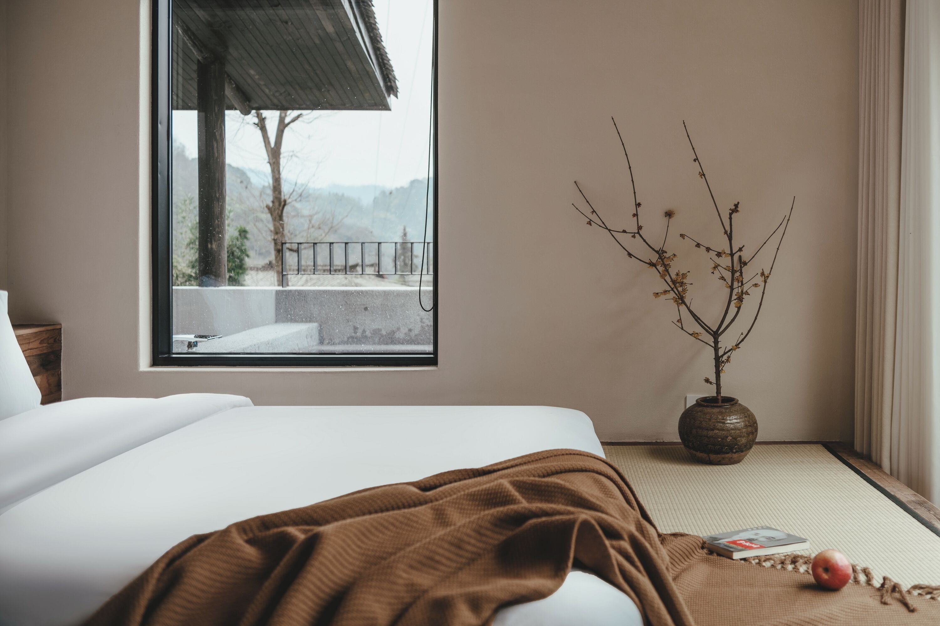 Une chambre avec une grande fenêtre, un lit, un vase avec des branches, et un livre et une pomme posés sur le sol.