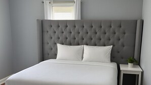 A modern bedroom with a grey tufted headboard, a white bedspread, and a small bedside table with a plant. - Heated Winter Stay • 3BR Home • Pet-Friendly • Free Parking • Near Hospitals • 6 (Manchester)