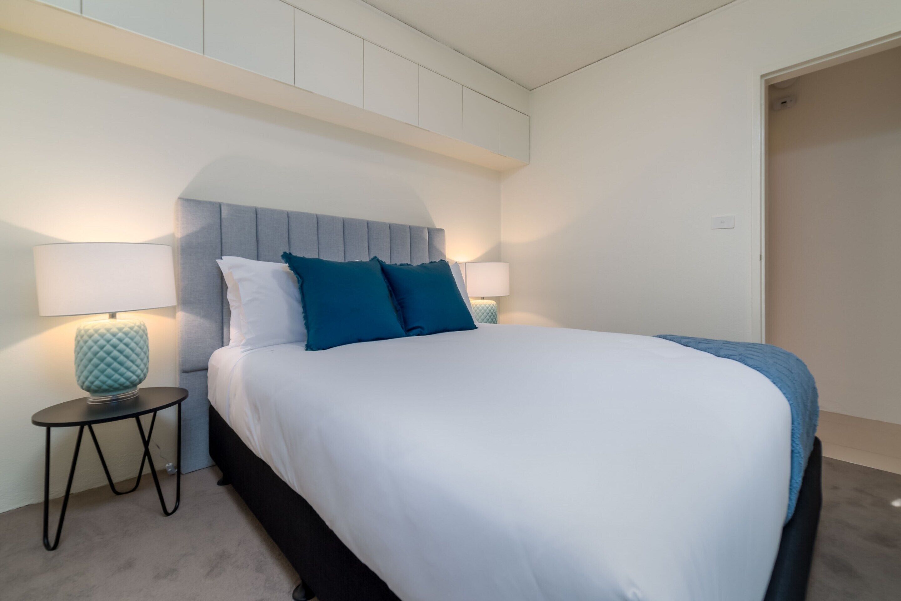 A neatly made bed with a grey headboard, two blue pillows, and a white bedspread. There is a small round table with a blue lamp beside the bed.