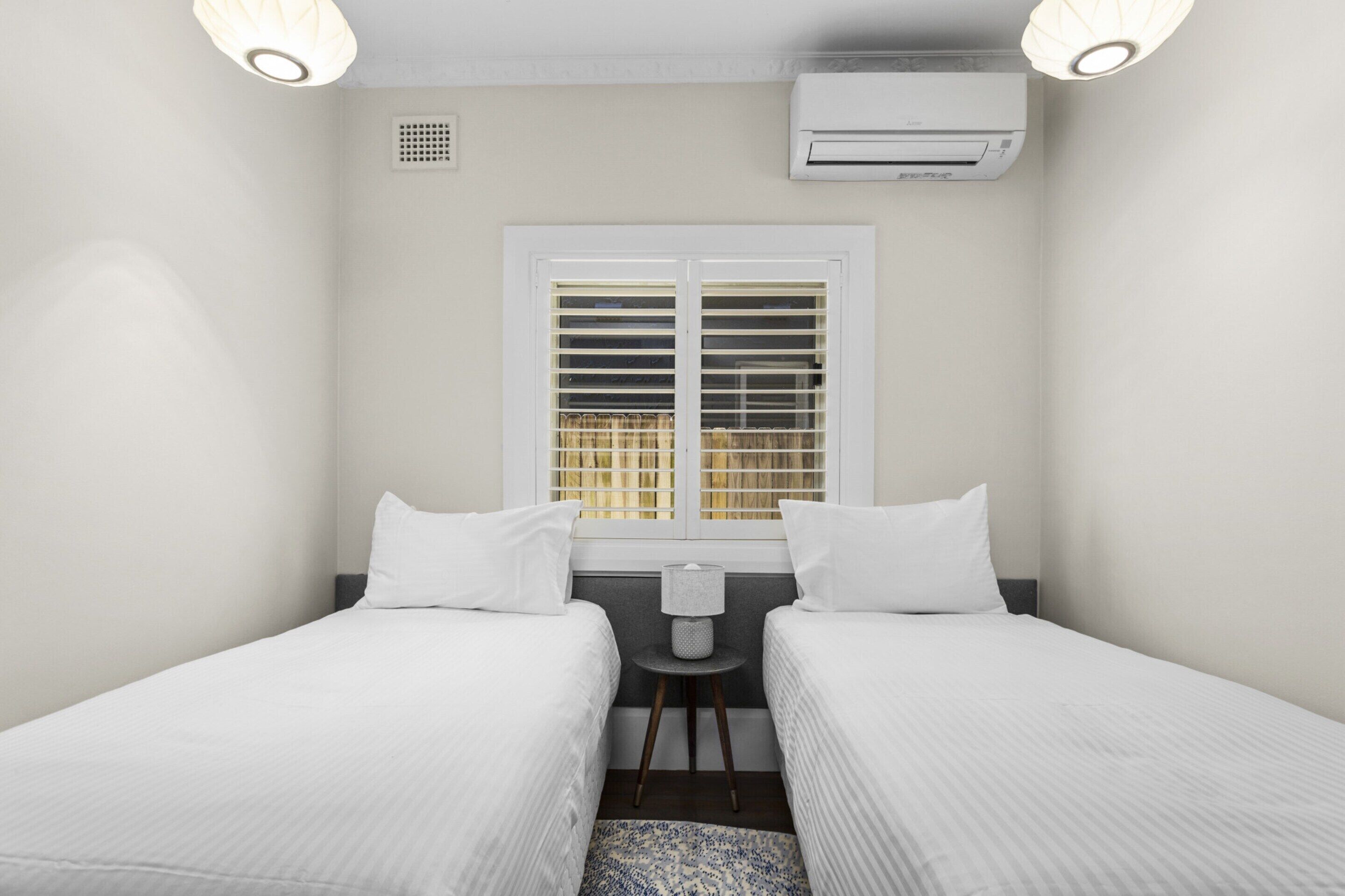 A small, clean room with two beds, a desk, and a window with blinds.
