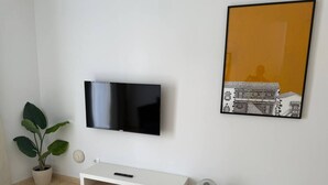 A room with a flat-screen TV mounted on the wall, a white TV stand with a remote control, a potted plant, a small white fan, and a framed picture of a building. - Don Diego - 2 bedroom in front of the beach (Playa De Los Cristianos)