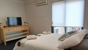 A bedroom with a bed, a TV on a wooden dresser, a window with blinds, and a balcony. - Spacious 2-bed Apartment in Heart of Leura (Leura)