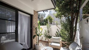Une terrasse ensoleillée avec une porte-fenêtre coulissante, une petite table avec un plateau, un banc rembourré et des plantes en pot.