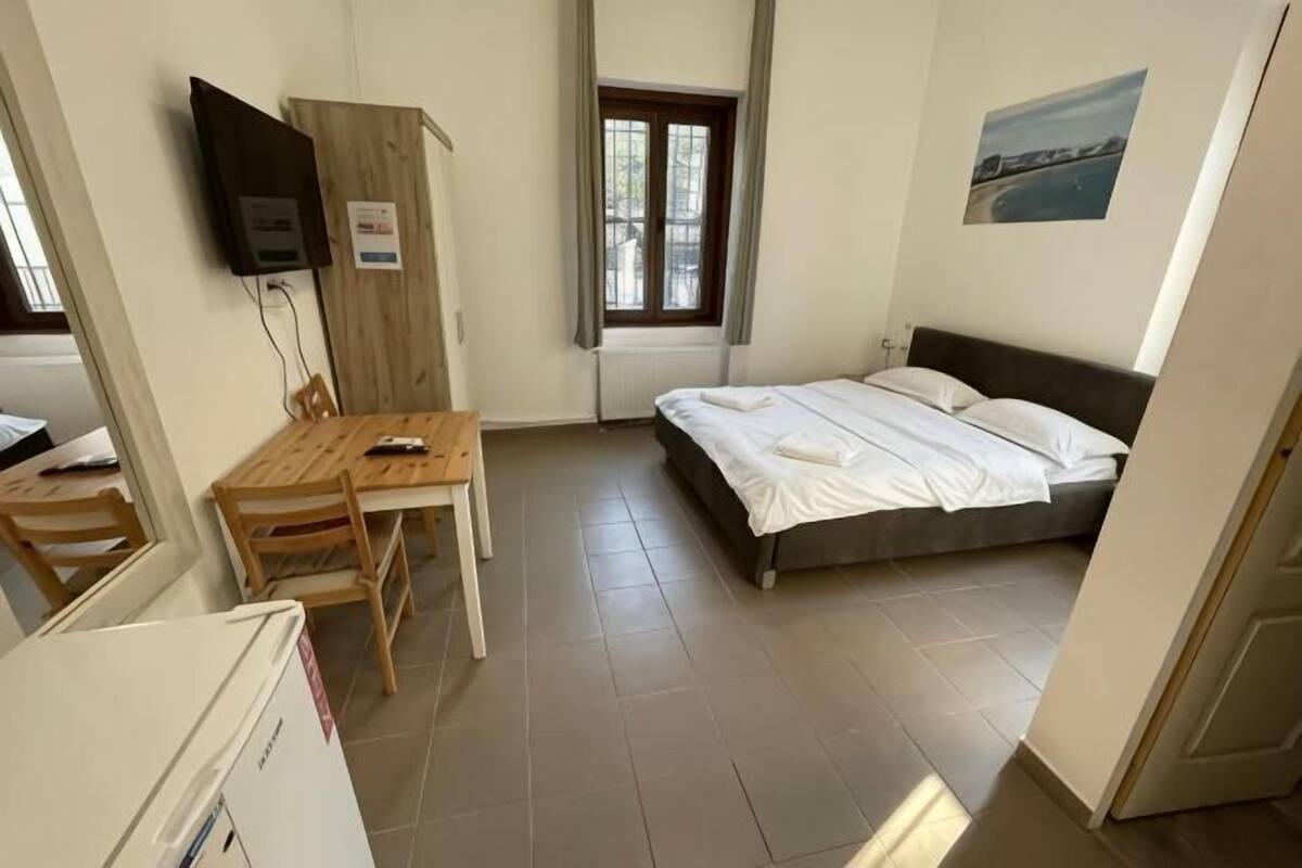A bedroom with a bed, a desk, a chair, a TV mounted on the wall, and a window.