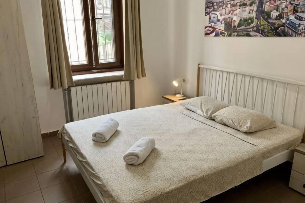 A neatly made bed with pillows and towels, a window with curtains, a nightstand with a lamp, and a cityscape painting on the wall.