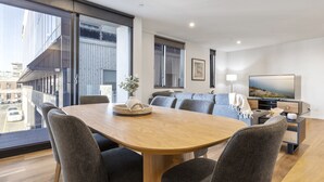 A modern dining area with a wooden table and grey upholstered chairs, large windows offering a view of the city, and a flat-screen TV on a stand. - Oceanfront 2-bed With Balcony & Study Nook (Geelong)