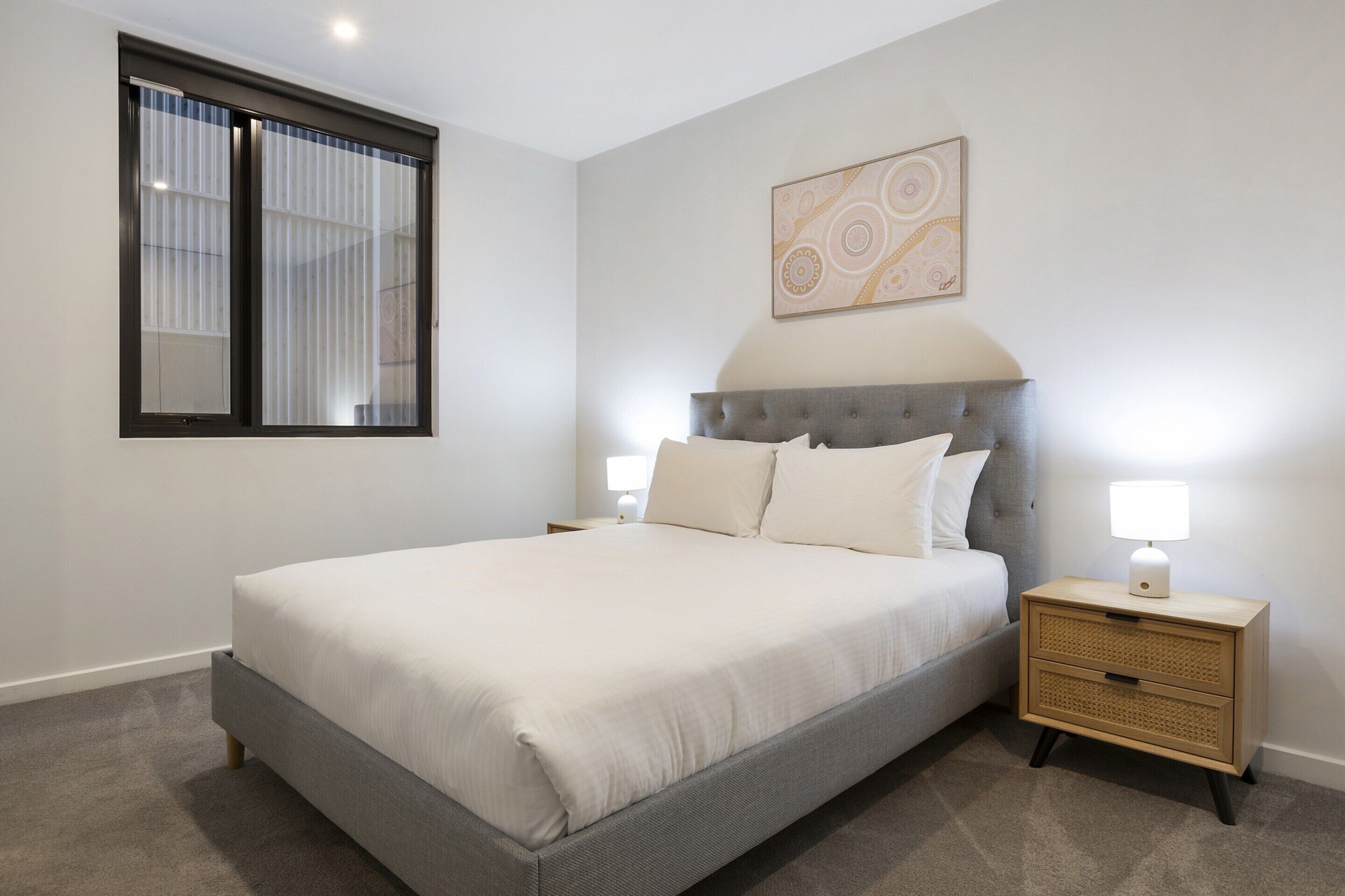 Ein Schlafzimmer mit einem großen Bett, zwei Nachttischen, einem Fenster mit Jalousien und einem an der Wand hängenden Kunstwerk.