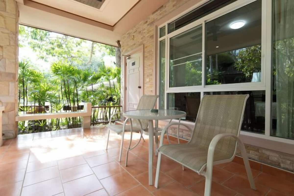 A covered patio with a table and chairs, a stone wall, and a view of greenery outside.