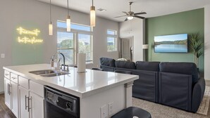 A modern kitchen with a central island, a dining area with a dark sofa, and a large window with blinds. - Charming 2-bedroom apartment with Pool, Gym. Close to Dwtn & Airport (Austin)