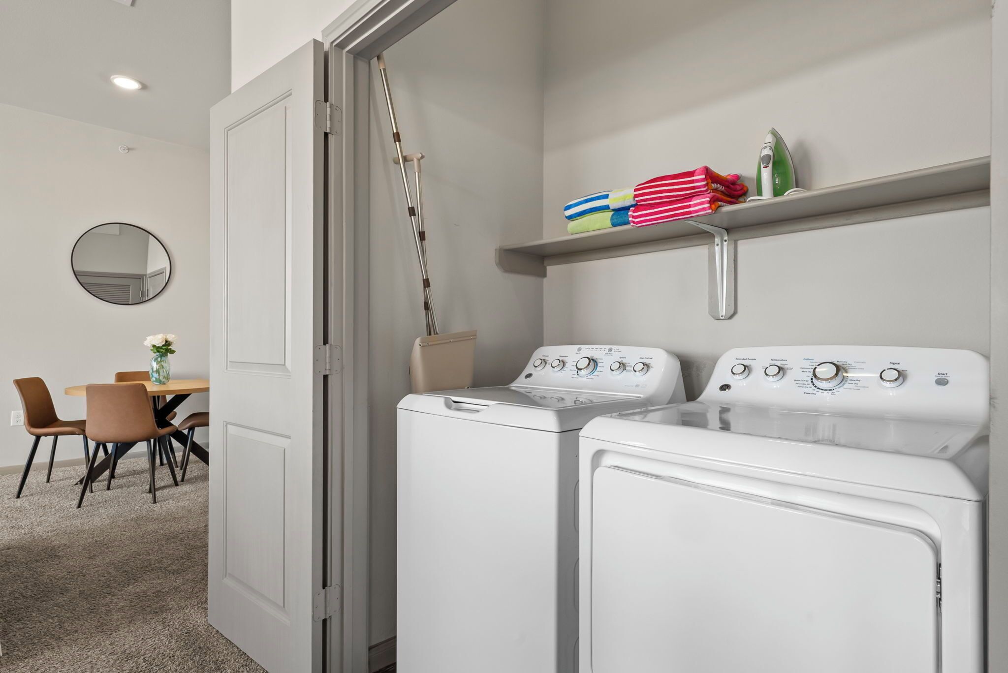 A laundry room with two white front-loading washers and dryers, a mirror on the wall, a dining area with a table and chairs, and a shelf with folded towels.