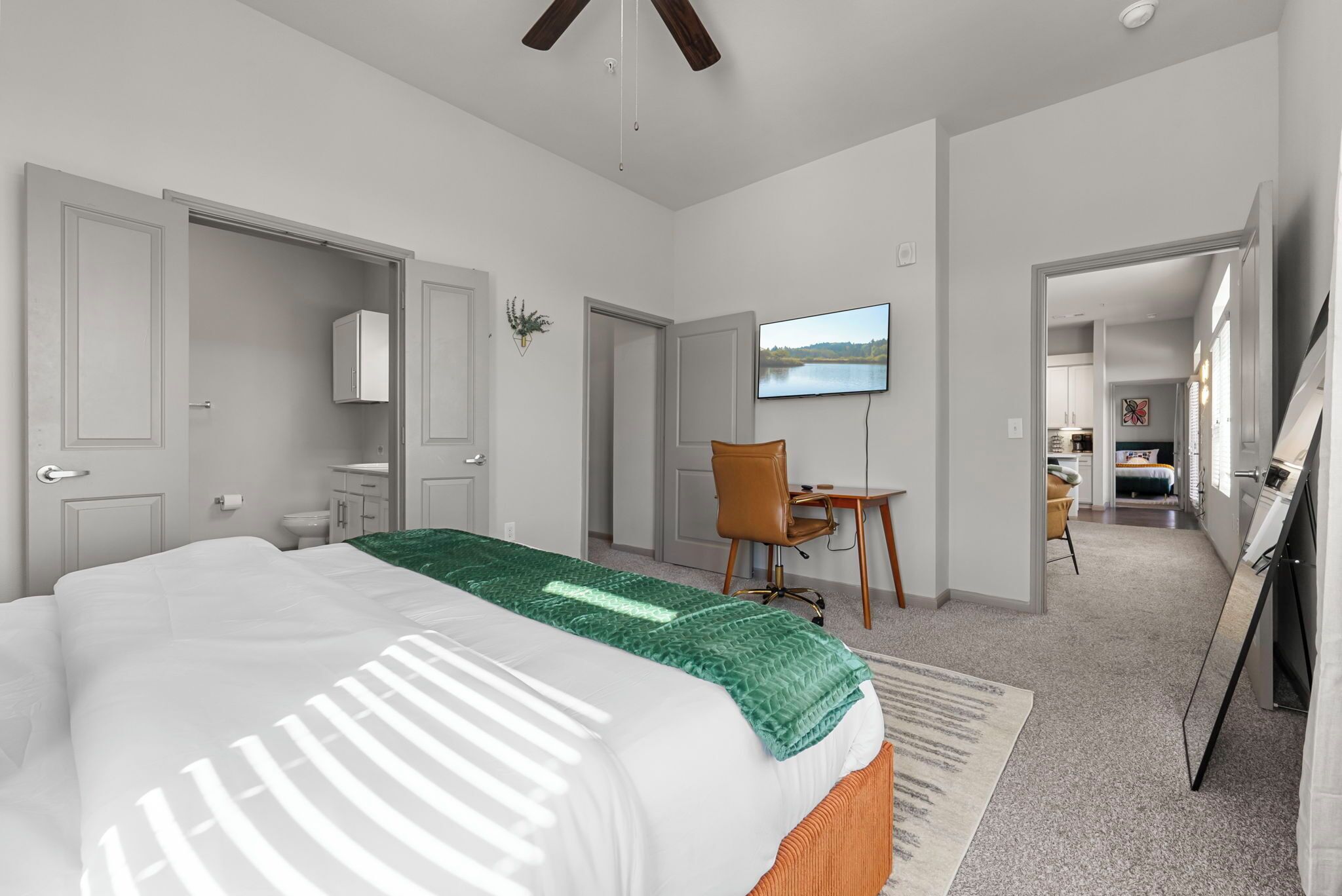 A modern hotel room with a large bed, a desk, a chair, a television, and a view of the kitchen and living area.