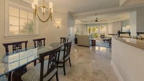 A spacious dining area with a glass table and chairs, a chandelier, and a view of the living room.