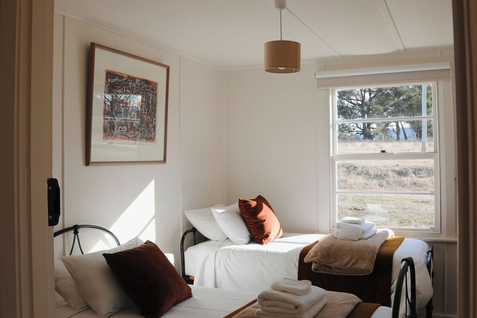 A room with a bed, a window with a view of trees, a hanging light fixture, and a framed picture on the wall.