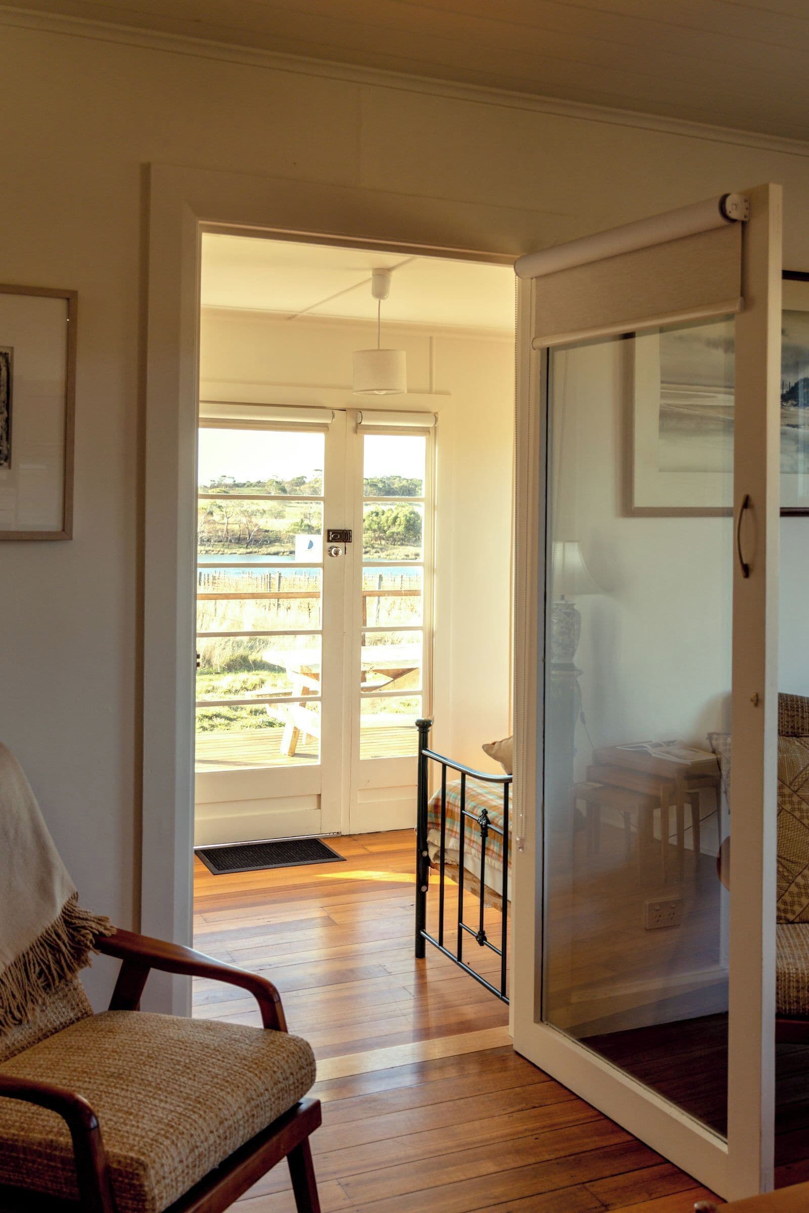 A room with a wooden floor, a chair, a bed with a patterned bedspread, a door leading to a balcony with a view of greenery and a body of water.