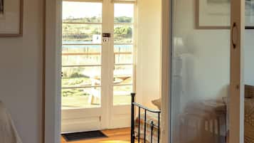 A room with a wooden floor, a chair, a bed with a patterned bedspread, a door leading to a balcony with a view of greenery and a body of water.