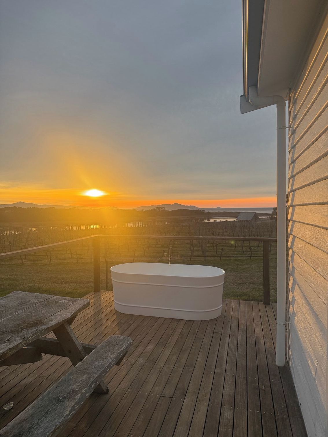 A wooden deck with a hot tub and picnic tables, overlooking a vineyard and a sunset.