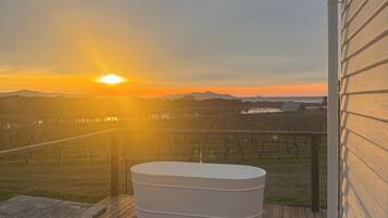 A wooden deck with a hot tub and picnic tables, overlooking a vineyard and a sunset.