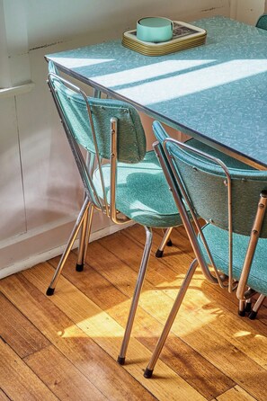A dining table with a teal tablecloth, two chairs with green upholstery, and a stack of plates on the table.