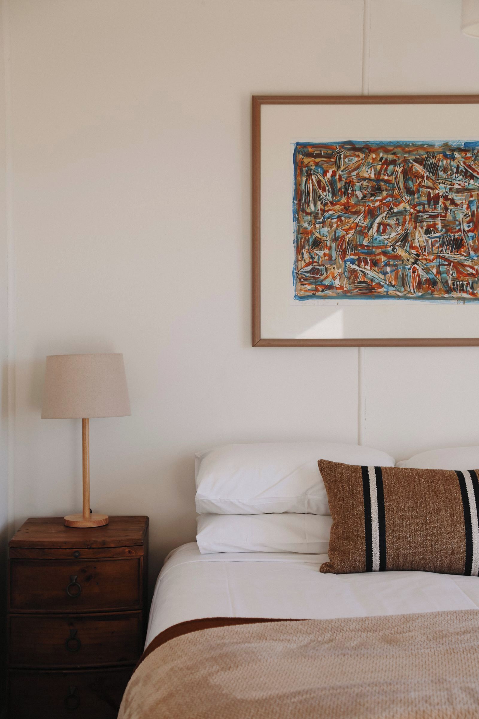 Een net opgemaakt bed met witte lakens en een bruin gestreept kussen, een houten nachtkastje met een lamp, en een lijst met een abstract schilderij aan de muur.