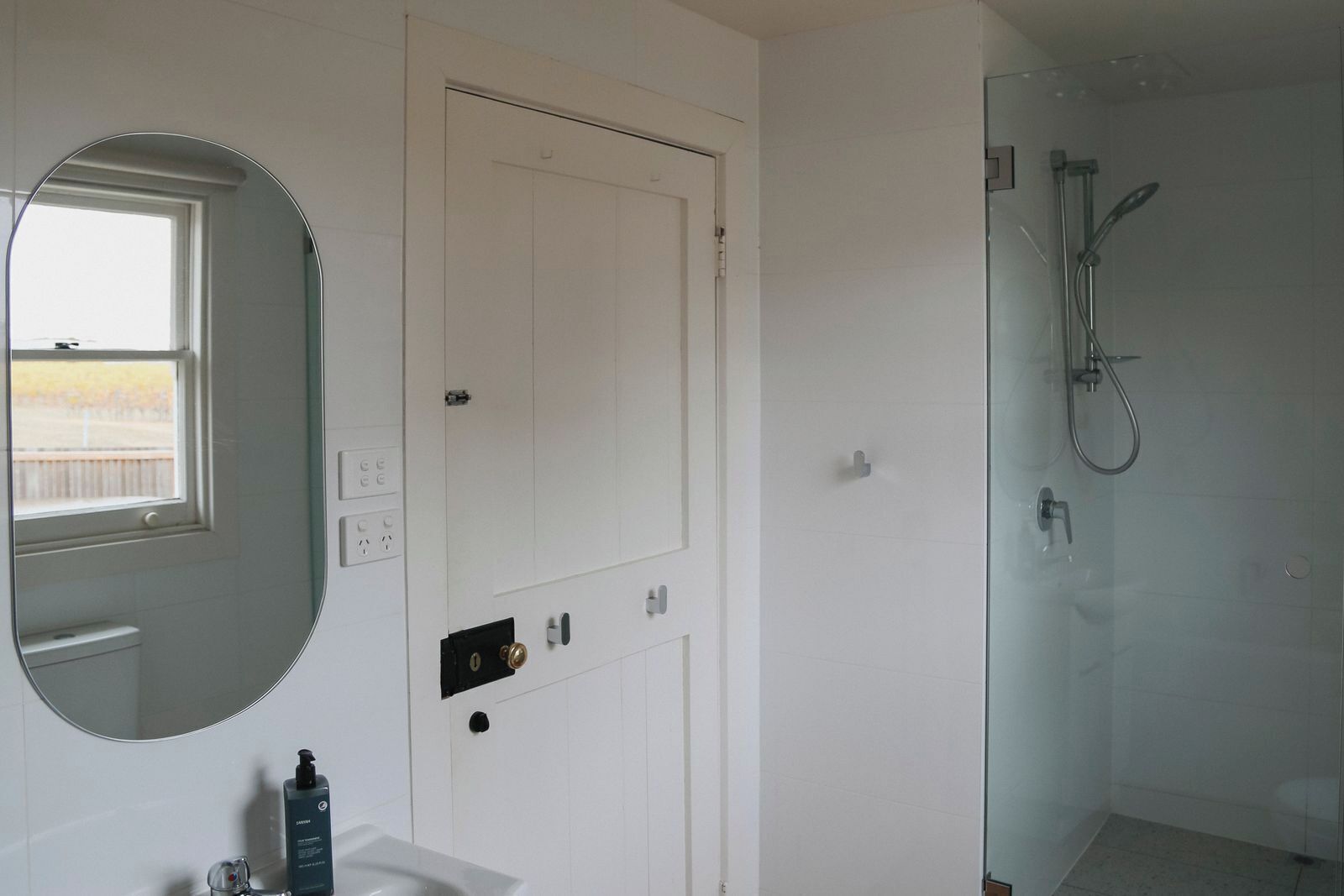 A bathroom with a white door, a round mirror, a glass shower enclosure, and a window with a view.