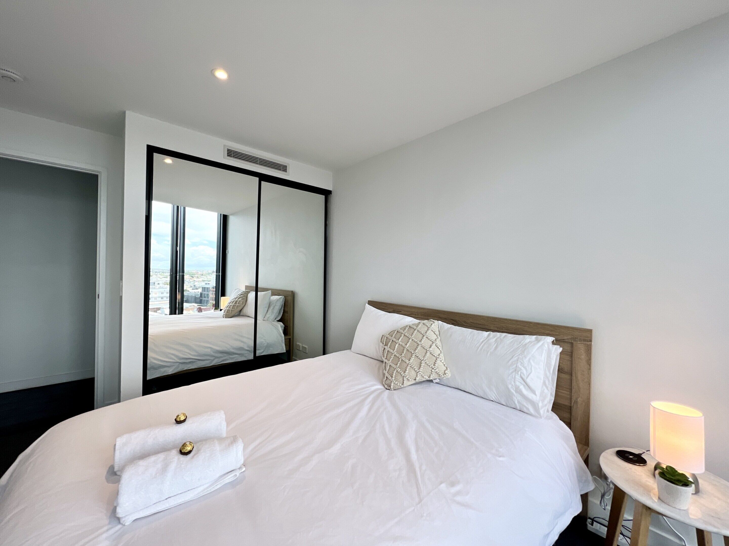 A modern hotel room with a large bed, a mirror, a bedside table with a lamp, and a view of the city through the window.