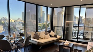 A modern living room with a sofa, dining table, chairs, and a view of a cityscape through large windows. - Stylish 1-bed With Pool, Gym & Free Parking (Brisbane)