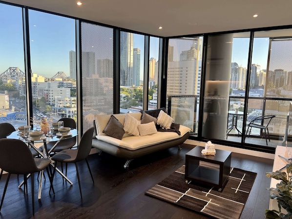 A modern living room with a sofa, dining table, chairs, and a view of a cityscape through large windows. - Stylish 1-bed With Pool, Gym & Free Parking (Brisbane)