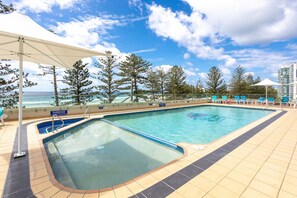 A rectangular swimming pool with a tiled deck, surrounded by lounge chairs and umbrellas, with a view of the ocean and trees in the background. - Beachfront 2-bed With Pool, BBQ (Gold Coast)