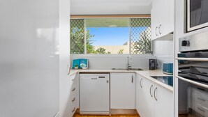 Cocina compacta con gabinetes blancos, lavavajillas, microondas y ventana con vistas a zonas verdes y edificios.
