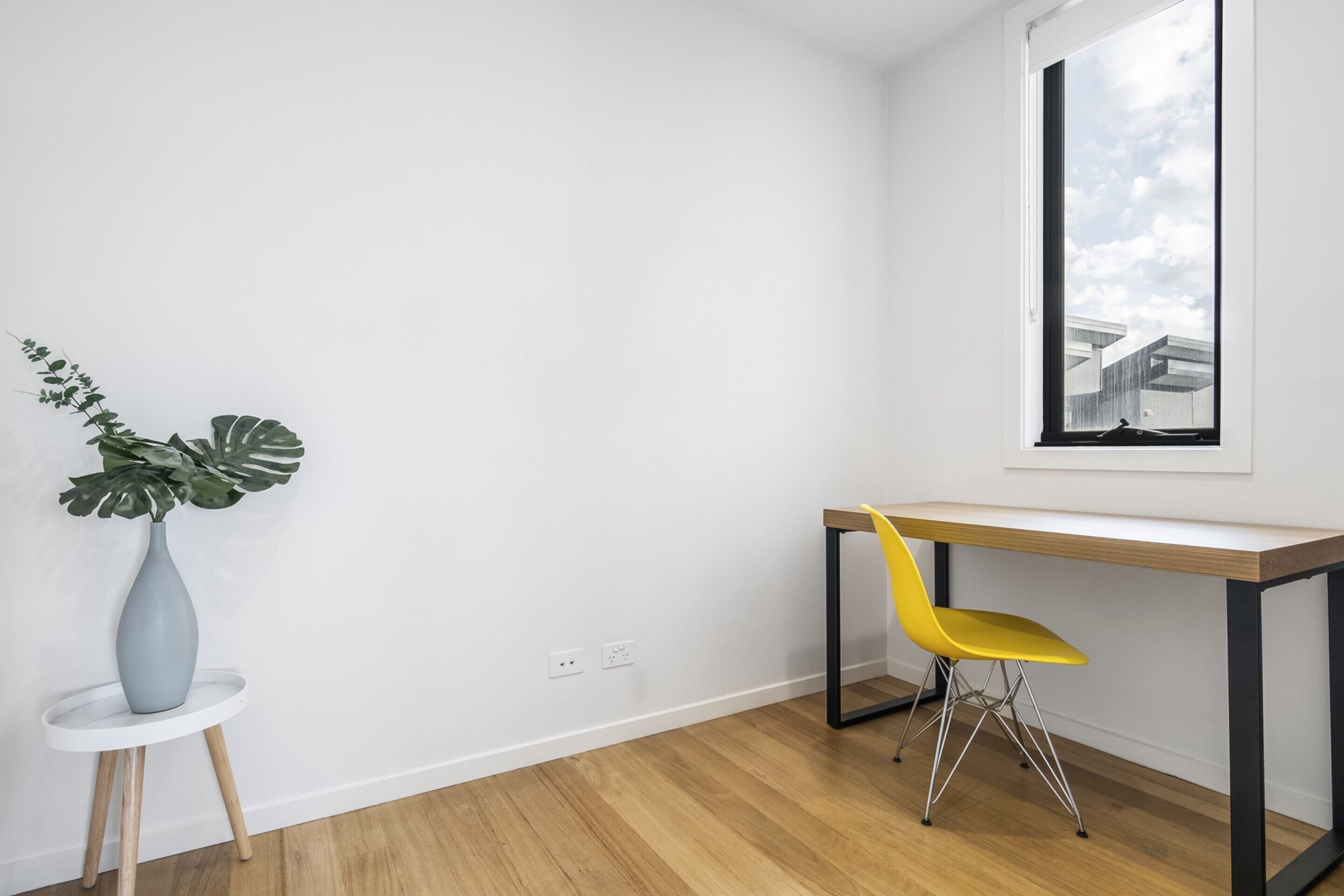 Une pièce avec un plancher en bois, un mur blanc, une fenêtre donnant sur des immeubles, une chaise jaune et un vase gris contenant une plante.