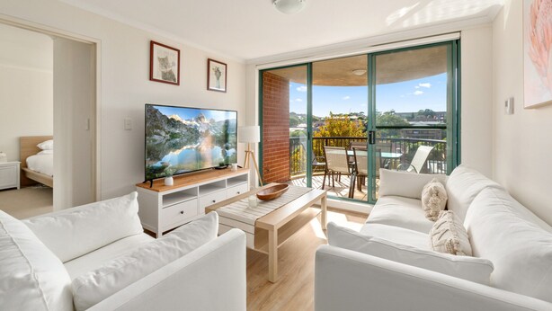 A modern living room with a flat-screen TV, white sofas, a wooden coffee table, and a balcony with a view. - Spacious 3-bed With Alfresco Dining by Amenities (Sydney)
