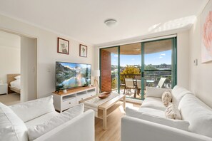 A modern living room with a flat-screen TV, white sofas, a wooden coffee table, and a balcony with a view. - Spacious 3-bed With Alfresco Dining by Amenities (Sydney)
