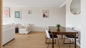 A modern living room with a dining area, featuring a white sofa, a small round table with a plant, and a wall-mounted mirror. - Spacious 3-bed With Alfresco Dining by Amenities (Sydney)