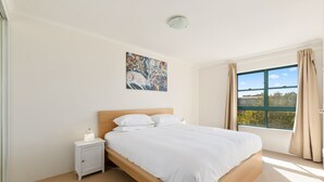 A bedroom with a large bed, a nightstand, a wall-mounted painting, and a window with curtains. - Spacious 3-bed With Alfresco Dining by Amenities (Sydney)