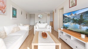 A modern living room with a white sofa, a wooden coffee table, a flat-screen TV, and a large window. - Spacious 3-bed With Alfresco Dining by Amenities (Sydney)