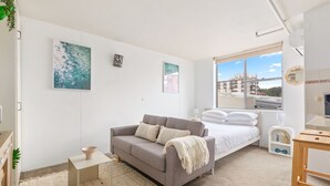 A modern bedroom with a grey sofa, a bed with white bedding, a small bedside table, and a window with a city view. - Bondi Studio Walk to Beach & Cafes (Sydney)