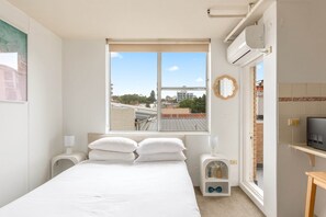 A bedroom with a large window, a bed with white linens, two bedside tables with lamps, a small shelf with a decorative item, and a TV on a stand. - Bondi Studio Walk to Beach & Cafes (Sydney)