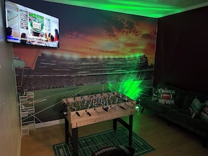 Een kamer met een groot muurschildering van een voetbalstadion, een tafelvoetbaltafel en een televisie waarop een sportevenement te zien is.
