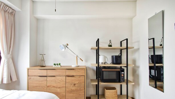 A bedroom with a bed, wooden dresser, shelves with kitchen appliances, and a mirror.