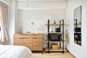 A bedroom with a bed, wooden dresser, shelves with kitchen appliances, and a mirror. - Modern Living Komagome 503, a compact studio at an excellent location (豊島区駒)