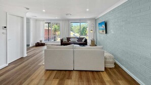 Un salon moderne avec un grand canapé gris, un téléviseur fixé au mur et une vue sur l’extérieur grâce à de grandes fenêtres.