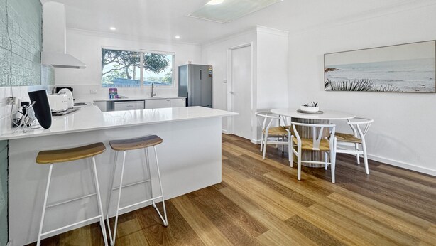Une cuisine moderne avec des meubles blancs, un îlot central doté de tabourets, un coin repas avec des chaises blanches et une grande fenêtre offrant une vue sur les arbres.