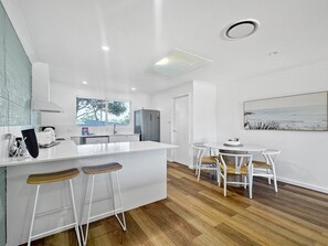 A modern kitchen with white cabinetry, a central island with stools, a dining area with white chairs, and a large window offering a view of trees. - Spacious 2-bed Townhouse With BBQ (Surf Beach)